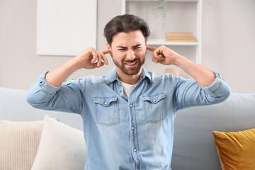 Young man with hearing problem covering his ears on sofa at home