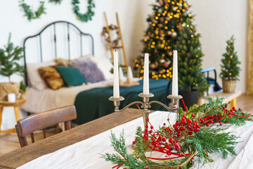 Three candles in candelabra on table surrounded by red berries and greenery on Christmas tree glows with warm lights background. Festive bedroom interior decorated for winter holidays