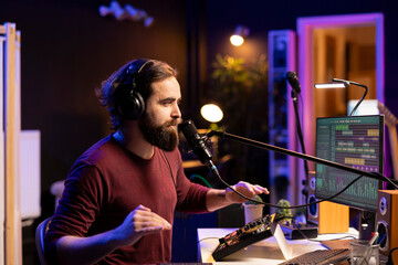 Young songwriter producer singing verses on microphone and adjusting volume levels on the melody,...