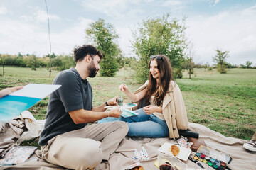 A young couple shares a joyful moment during a picnic in a lush park. Surrounded by nature, they enjoy food and drinks while engaging in creative activities on a blanket.