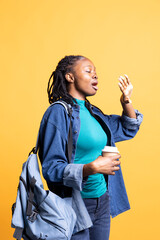 Young girl with backpack yawning, feeling tired, drinking coffee to wake up on her way to school. Fatigued student using caffeinated beverage to gain energy, lacking sleep, studio background