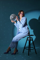 Beautiful young woman in stylish outfit sitting with disco ball on stool against blue background