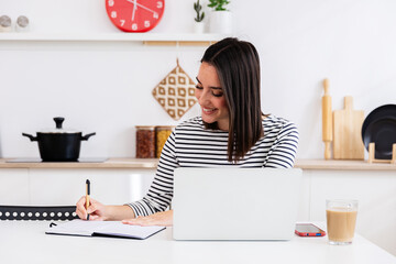 Young adult student woman taking notes while using laptop computer in the kitchen. Millennial...