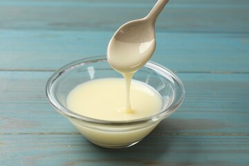 Condensed milk flowing down from spoon into bowl on light blue wooden table, closeup