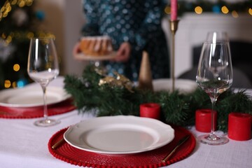 Woman setting table for Christmas dinner at home, focus on plate, glasses and decor