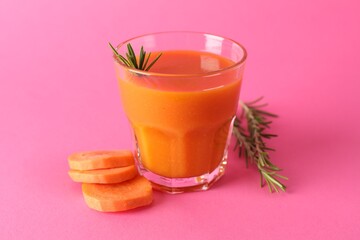 Fresh carrot smoothie in glass, rosemary and cut vegetable on pink background, closeup