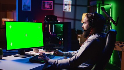Man in dimly lit apartment playing video games on isolated screen gaming PC at computer desk, enjoying day off from work. Gamer battling enemies in online multiplayer shooter on chroma key monitor