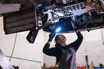 Female chinese mechanic inspecting car engine with flashlight in repair shop