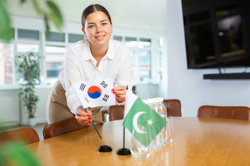 Positive young woman putting little flag of South Korea on table next to the flag of Pakistan and...