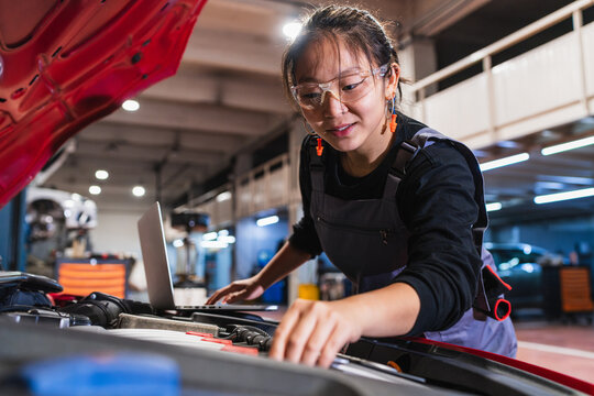 Young chinese female mechanic inspecting car engine with laptop in repair shop