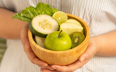 Close-up of hands holding a wooden bowl filled with fresh green apple, cucumber, lime, kiwi, and lettuce, highlighting vibrant natural colors.
