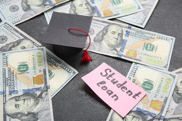 Paper sheet with text STUDENT LOAN, dollar banknotes and small graduation hat on dark background
