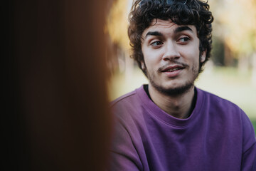 A young man wearing a purple sweater enjoys a casual conversation outdoors. The natural background and relaxed expression capture a moment of informal interaction and leisure in the park.