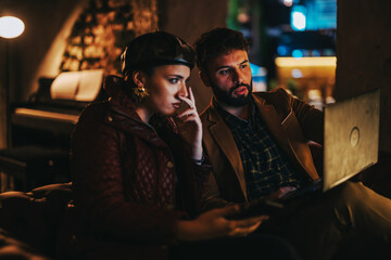 A man and woman focus intently on a laptop screen in a dimly lit room, conveying teamwork and concentration. Their expressions suggest deep engagement in a nighttime work session.