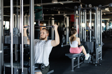 Fototapeta premium Young athletic man in sportswear training his arms muscles on machine in gym