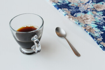 Transparent glass coffee cup and silver spoon on white table with blue napkin on the right top corner. Hot soft drink, caffeine, stimulating beverage.