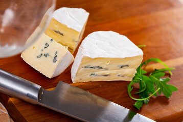 Pieces of piquant soft blue cheese lying on wooden cutting board with fresh green arugula and kitchen knife. Artisanal dairy product