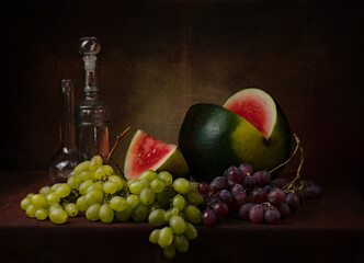 still life with a cut red watermelon with different varieties of green and black grapes and a couple of decorative bottles on the table on a dark background