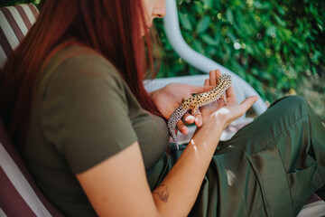 A girl sits outdoors, playing gently with a spotted lizard on her hands, surrounded by nature. Her calm demeanor and closeness to the reptile create an atmosphere of curiosity and tranquility.