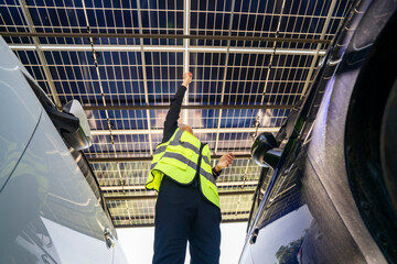 Engineer inspecting solar panels in a solar-powered carport surrounded by parked cars. Renewable...