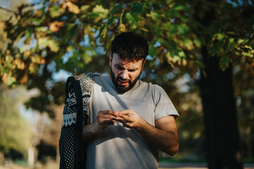 Obraz premium A man stands under a tree with colorful autumn leaves, closely examining something small in his hands. The sunlight filters through the branches, creating a serene and contemplative atmosphere.