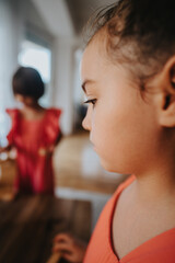 Close-up of a pensive child indoors, with another child in the background. The image conveys innocence, imagination, and childhood moments. Captured in a bright home environment.