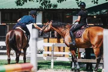 Two equestrians are seen interacting with their horses in a stable. They are wearing riding gear and appear to be preparing for a ride. The scene conveys teamwork and companionship.