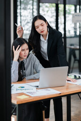 Two professional women in business attire working together at a desk with a laptop and charts in a contemporary office.