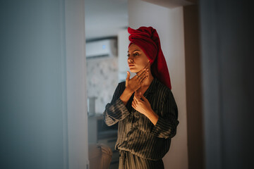 A woman stands in front of a bathroom mirror, wrapped in a red towel, applying skincare. She...
