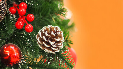 Close-up of a Christmas decoration featuring a frosted pinecone, red berries, and a shiny red ornament against green foliage and an orange background. Perfect for holiday-themed visuals.