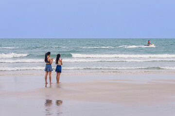 Back view of happy carefree women using mobile phone at beach. Woman friends using smartphone at seaside. Beautiful lovely two women spending time at beach. Concept of travel, tourism and vacation.
