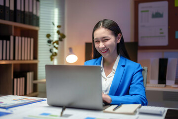 Happy businesswoman using laptop computer, working late in office with financial charts and office documents on desk