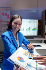 Two businesswomen analyzing financial charts and data in a brightly lit office at night, working...