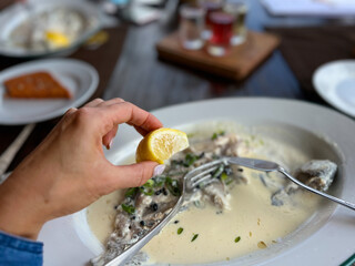 Golden trout in cream sauce in white plate at restaurant. Hand holding a slice of lemon on a plate with fish and tableware.