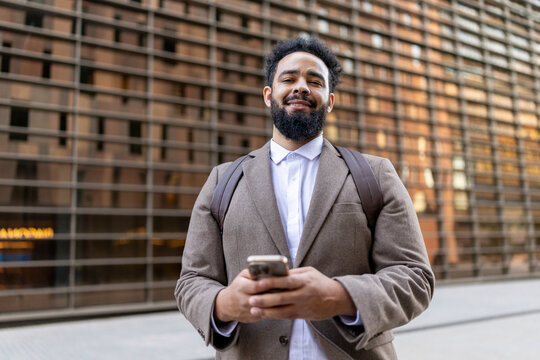A confident African American businessman stands outdoors, holding his smartphone. He wears a stylish brown suit and crisp white shirt, with a modern corporate building in the background