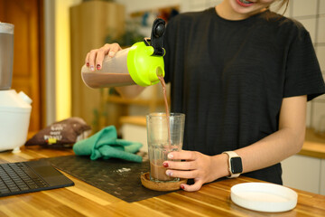 Smiling woman in sportswear pouring a freshly made chocolate protein shake into a glass