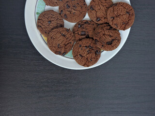 A top-down view of a plate filled with chocolate chip cookies on a dark wooden table. The cookies are rich brown with visible chocolate chips