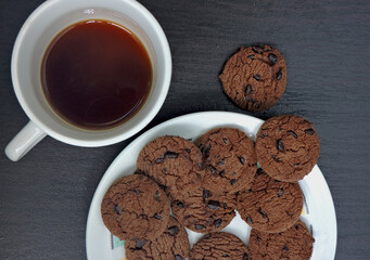 A top-down view of a plate filled with chocolate chip cookies on a dark wooden table. The cookies are rich brown with visible chocolate chips. Perfect for themes like food, snacks, desserts