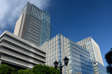 Obraz premium Modern skyscrapers with glass and beige facades rise against a deep blue sky in Roppongi, Tokyo. Green trees and decorative street lamps add contrast at ground level.