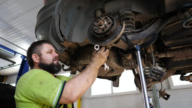 Male auto mechanic works underneath a lifting vehicle at workshop. Adult professional repairman fixing car at service or garage. Concept of regular automobile maintenance. Close up Slow motion