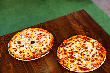 Two delicious pizzas with golden crust and melted cheese are placed on a rustic wooden table outdoors.