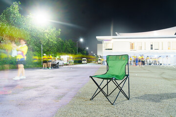green folding chair sits alone on well lit street at night, surrounded by blurred figures and...
