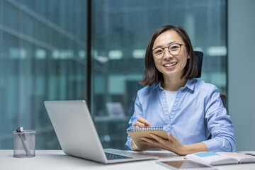 Asian woman businesswoman using a notepad and laptop, engaging in writing or planning in a professional office environment. The positive expression and productive atmosphere