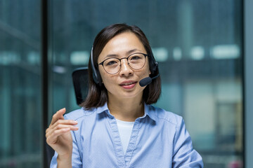 Asian woman wearing a headset engaging in a professional online meeting or video call. She is...
