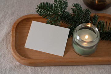 Blank paper card mockup, green candle with soft dim light, fir branch on wooden tray on table, cozy...