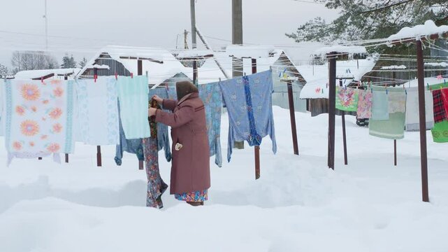 Traditional daily laundry far from civilization, Grandmother gray downy shawl, felt boots goes hang out laundry robe flowers. Clean sheets, freshness winter snow outback, wood village Russia, Siberia