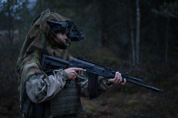 A soldier woman in camouflage, wearing a tactical helmet with a night vision monocular and an assault rifle in her hand in a dark forest.