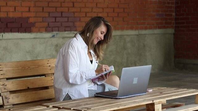 Smiling blond businesswoman in white blazer sitting on wooden pallets  using typing on laptop computer and have conversation online gesticulating, studying outdoors. Female psychologist working remote