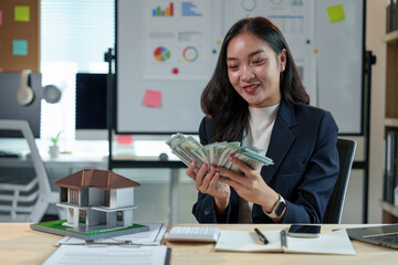 Asian sales girl, real estate agent holding money, counting money from house sale, customer's lease agreement, insurance while sitting at desk in office with model house in real estate project.