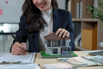 Asian saleswoman, real estate agent holding model house and writing lease contract, insurance,...